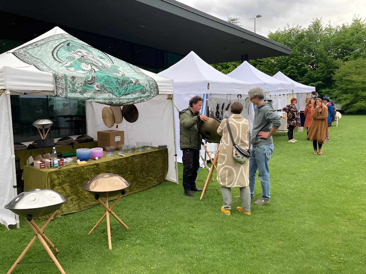 Festivalmarkt im Freien: Stände vor dem Gebäude, Handpans auf Ständern, Besucherinnen und Besucher im Gespräch.