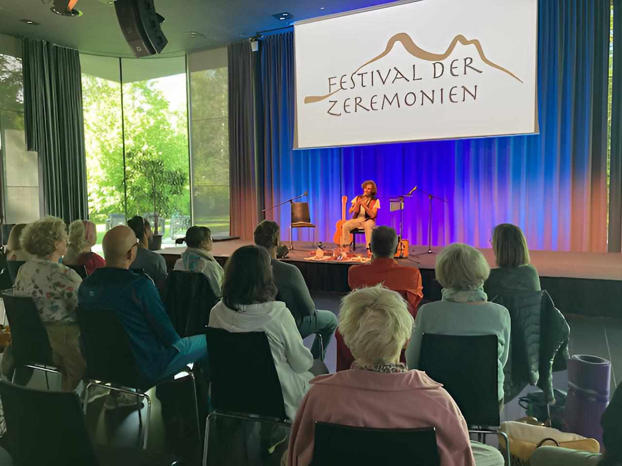 Publikum im Saal vor Bühne mit Musikerin, Banner ‚Festival der Zeremonien‘ im Hintergrund.