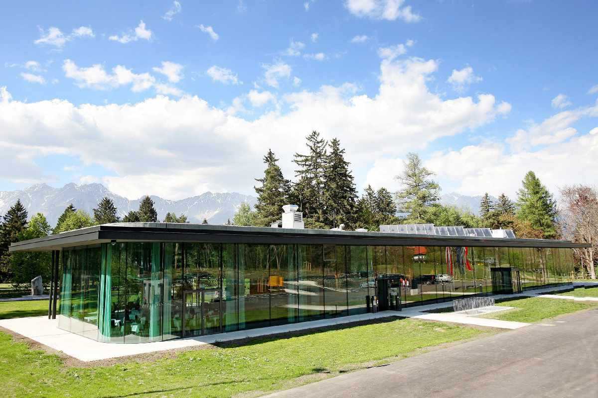 Weite Ansicht des Congresspark Igls: flaches Glasgebäude, Wiese und Berge im Hintergrund.“